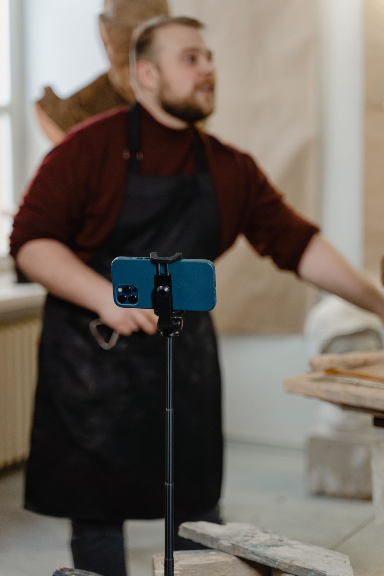 Celular no tripé, em primeiro plano, grava um artesão de avental na oficina