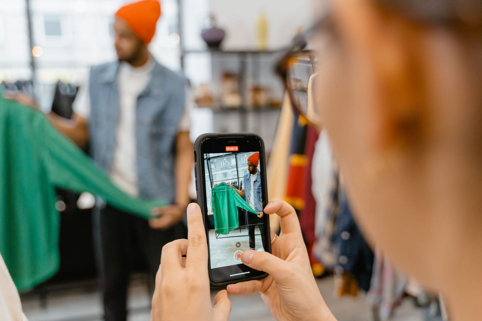Mãos seguram o celular que grava um vendedor de gorro laranja mostrando uma jaqueta verde na loja