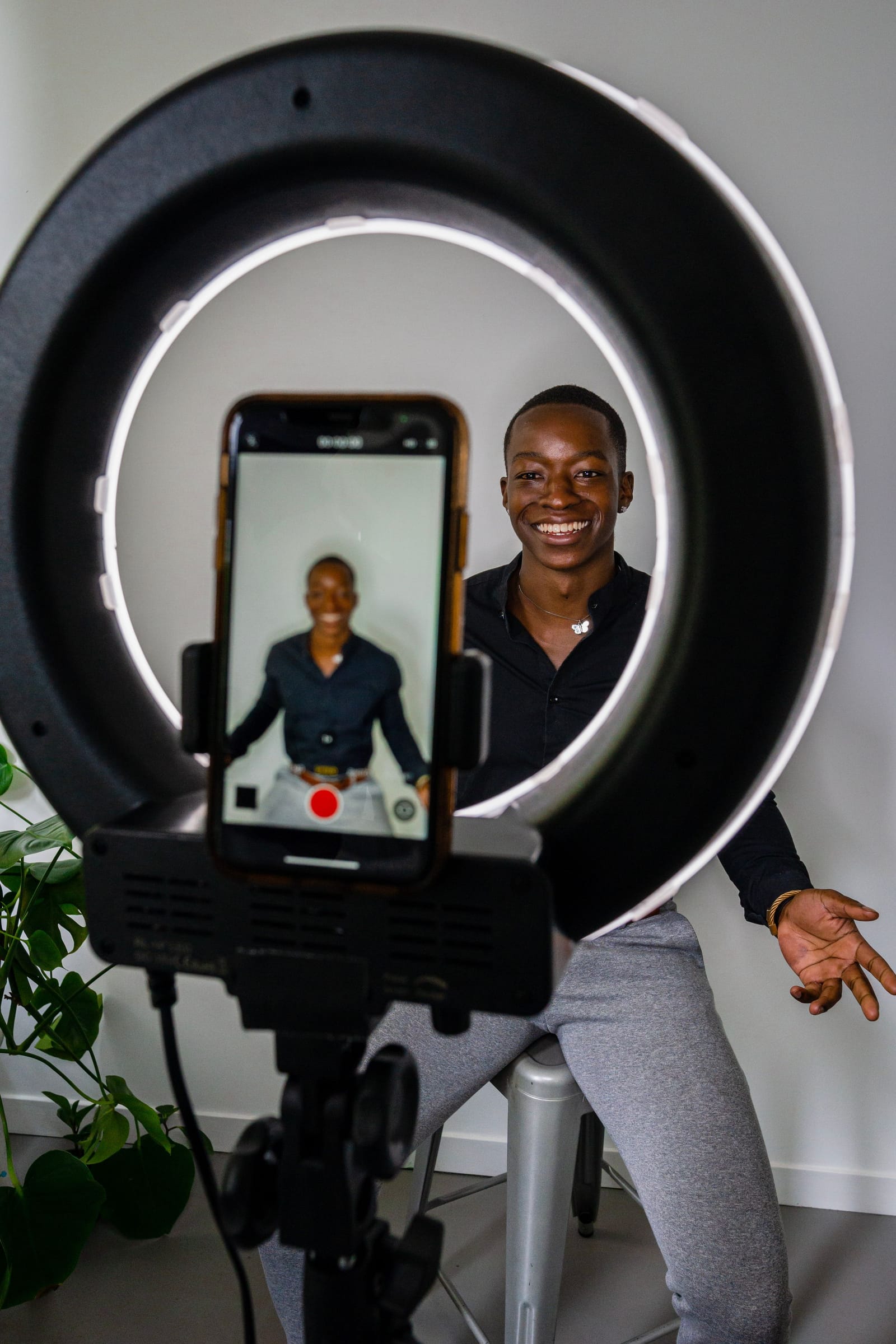 Homem sorridente sentado diante de um ring light grande; em primeiro plano, o celular o mostra na tela, pronto para gravar