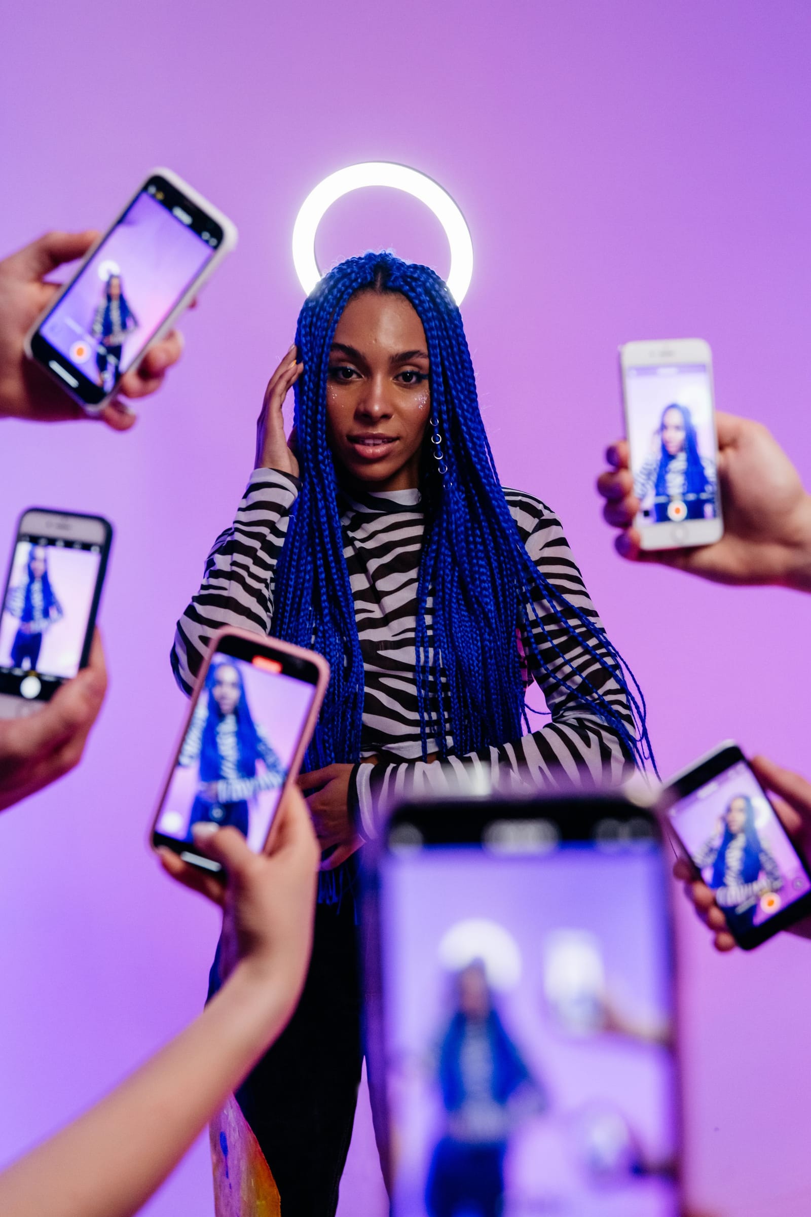 Criadora de tranças azuis posa diante do ring light, cercada por várias mãos que a filmam com celulares