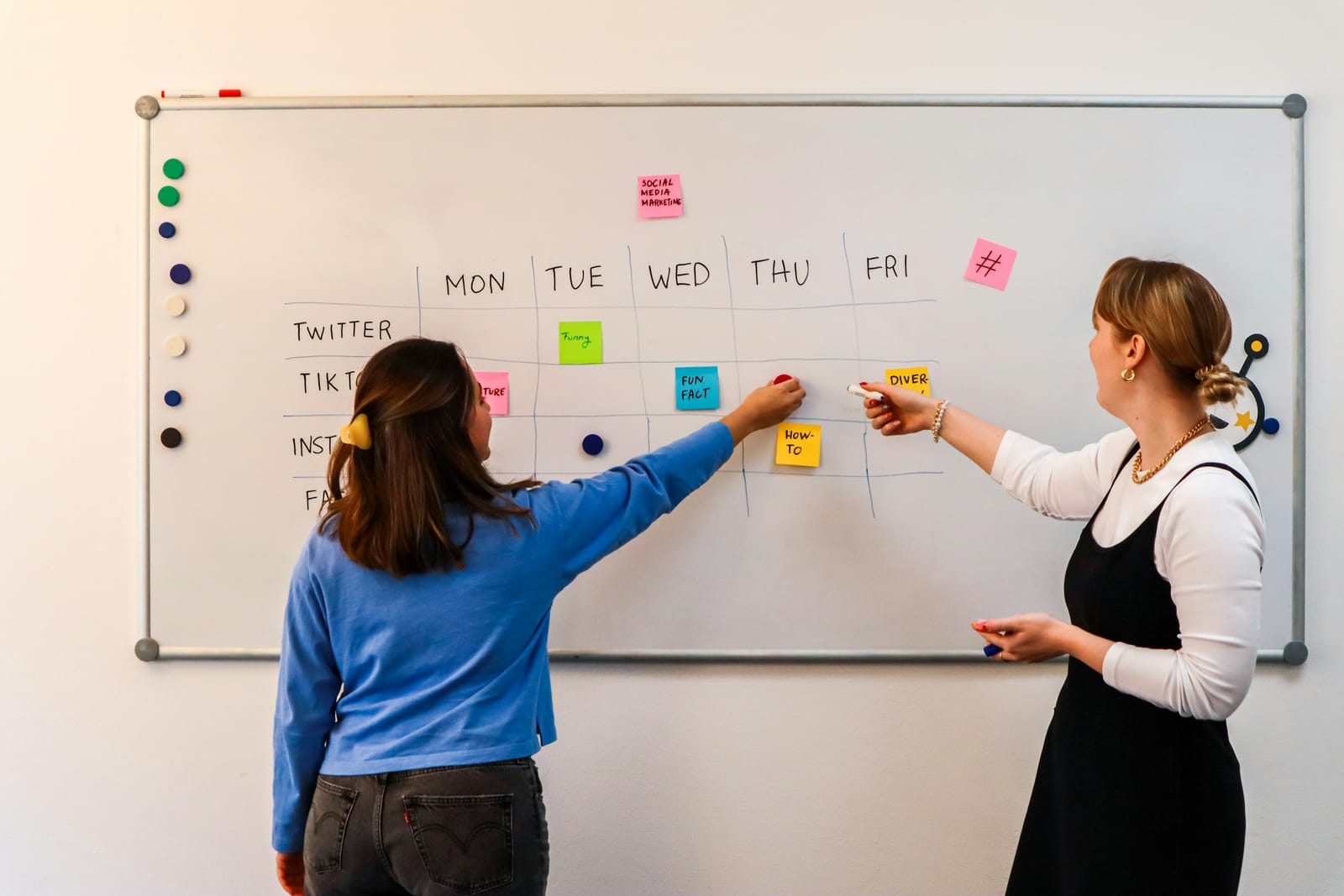 Duas mulheres montam no quadro branco o calendário semanal de conteúdo, de segunda a sexta, com post-its coloridos